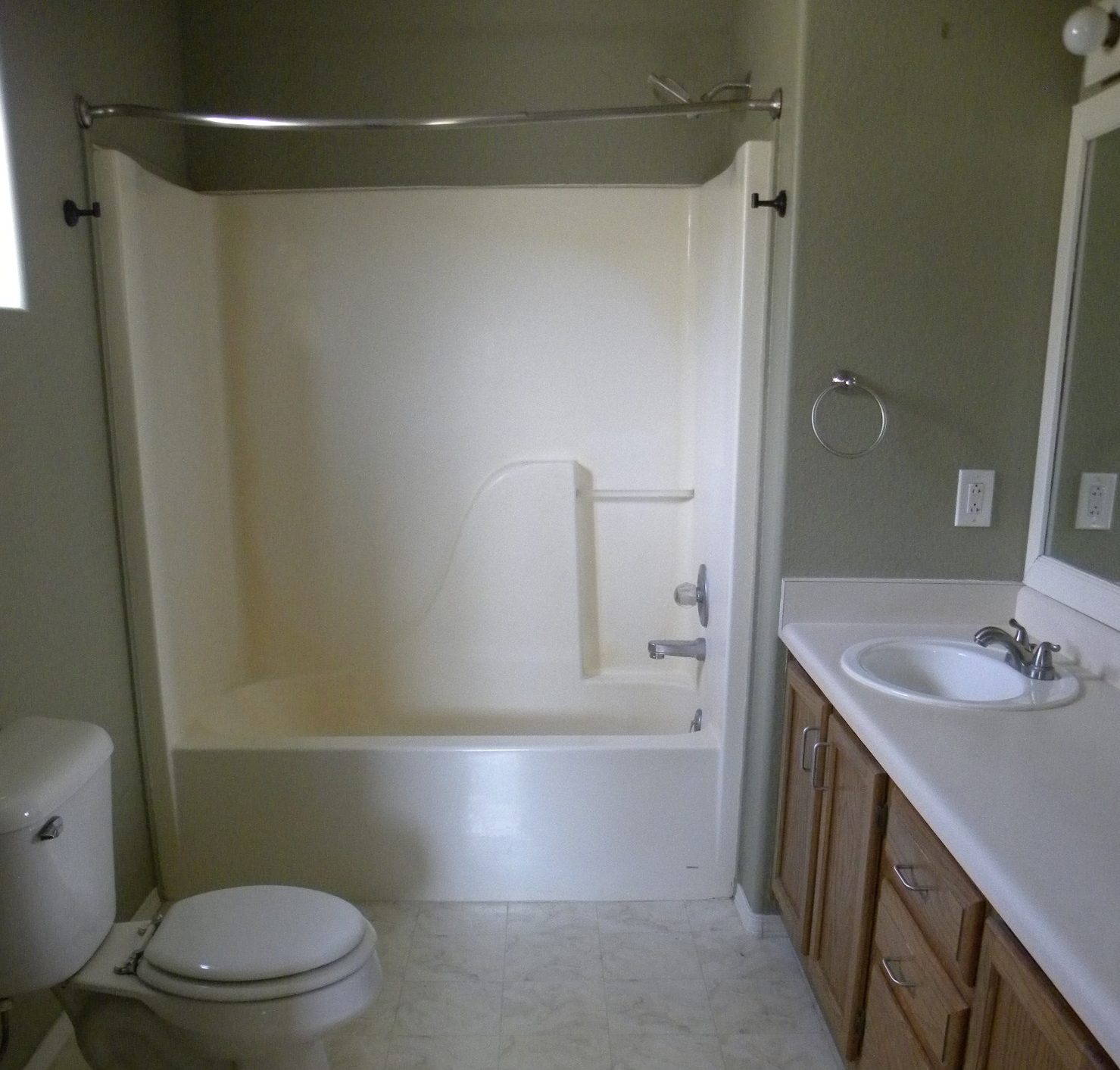 Before photo of a residential bathroom with white countertops and wooden cabinets, a white toilet, a white shower, and white marbled tiles.